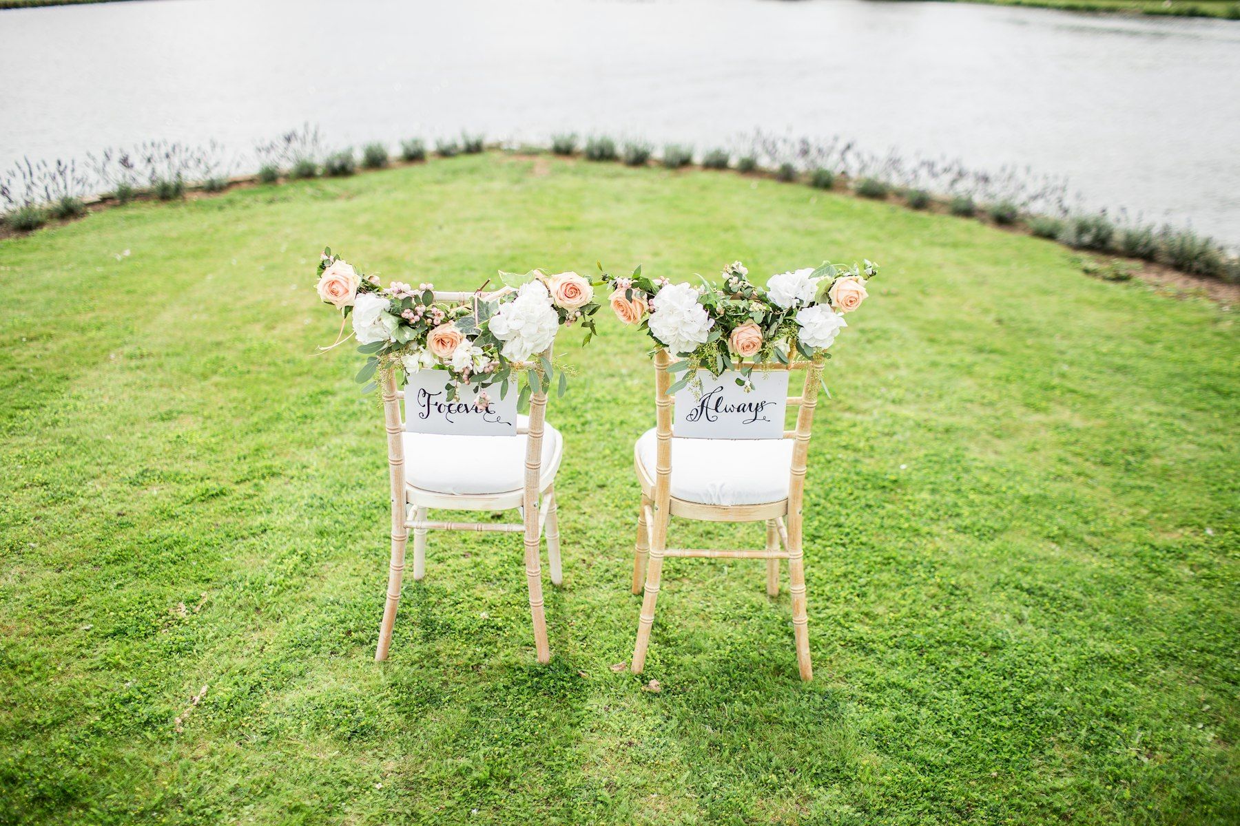 Two white chairs dressed with flowers on a lawn by the water