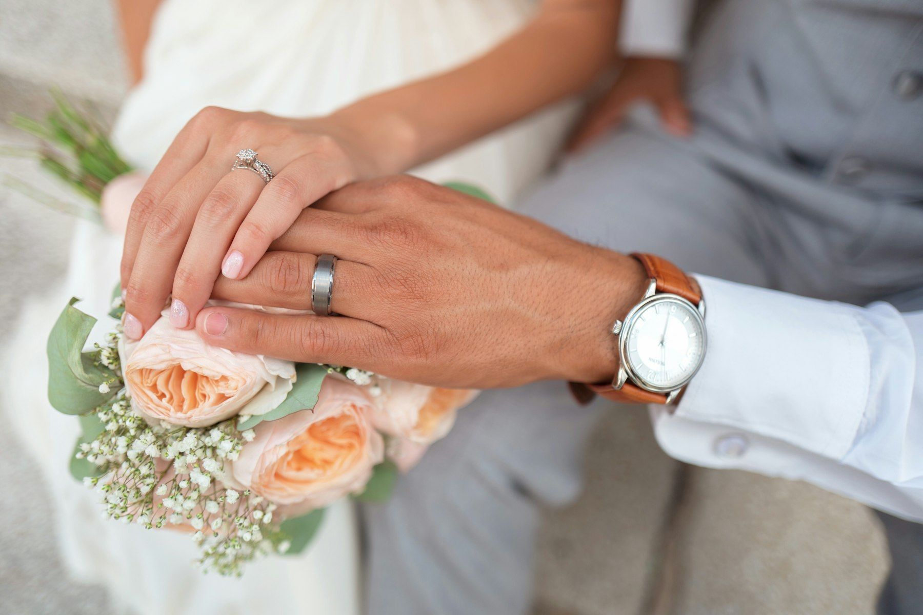 Two hands with wedding rings resting on a bouquet of peach roses