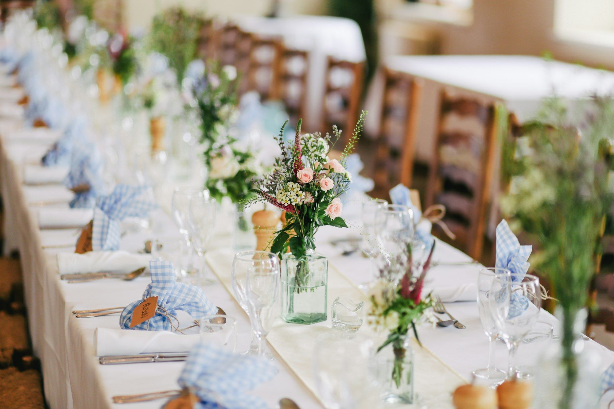 Long reception tables set with white linens, glassware and small flower arrangements